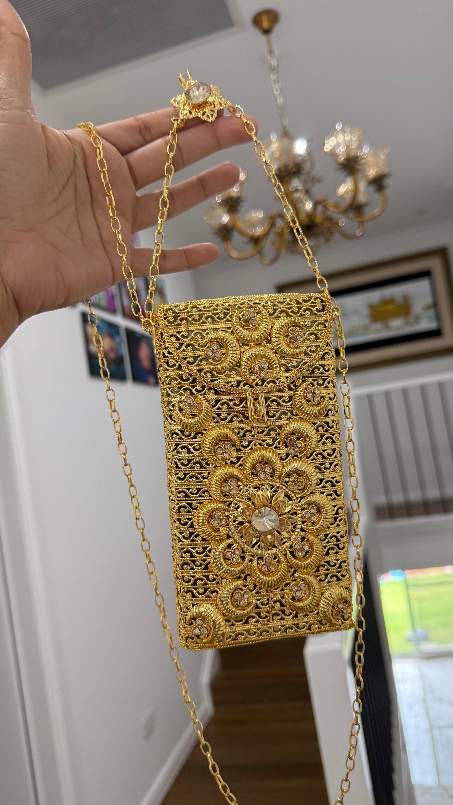 Gold lace handbag with chain strap held by a hand, indoor setting with chandelier.
